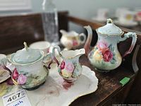 Hand painted teapot, creamer, and sugar bowl on wooden surface showing floral and gold details.