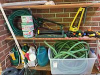 Shelf with coiled green garden hoses, rolls of plastic lawn edging, and assorted garden tools and watering cans.