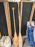 Photo showing two wood canoe paddles standing upright against a wall with some folded chairs nearby.
