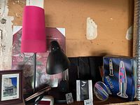 Photo showing a tall table lamp with bright pink shade, black desk lamp, Blob Lamp box, framed photos, and mirrors against a wooden background.