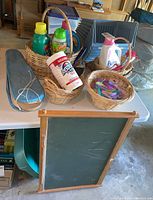 View of various laundry detergent bottles and fabric softeners in woven baskets, a container of stain remover, a laundry brush, and a woven basket with plastic clothes pins on a table, with a framed chalkboard displayed below.