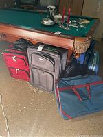 Six pieces of luggage including red, gray, navy blue, and hard shell light blue suitcase arranged by a pool table.