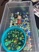 Plastic container filled with a large variety of glass marbles, including many different colors and sizes, with a blue bowl inside holding more marbles.