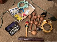 Various percussion instruments including tambourines, maracas, castanets, recorder, and a case with metal parts for instrument repair or assembly. Items are placed on carpeted floor.