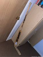 Full view of the wooden Hespeler goalie stick leaning against a wall and cabinet in a basement-like setting.