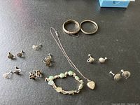 Full lot view showing two bangles, heart-shaped locket necklace, green and white stone bracelet, and six pairs of screw back earrings arranged on black surface
