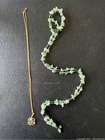 Two necklaces displayed side by side on black background: gold-tone chain with Swarovski clear crystal clover flower pendant, and long green semi-precious stone chip rope necklace with metallic hematite beads.