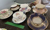 Six assorted porcelain teacups and saucers on dark table, various floral patterns and designs visible.