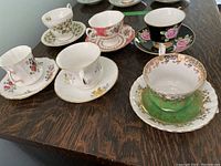 Top view showing all six decorative teacups with their matching saucers on a wooden table, showcasing variety of floral patterns and colors