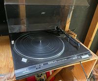 Front and angled view of black Technics SL-B20 turntable showing platter, tonearm with cartridge, and clear dust cover in open position on a wooden surface.