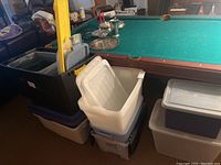 Black, dark blue, and clear plastic tote bins, some stacked on each other, near pool table.