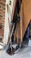 Photo showing long-handled garden tools including a metal garden fork, metal rake with fan tines, edger with metal blade, all with wooden handles, stored in a garage.