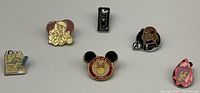 Six Disney and Pixar themed collectible pins displayed on fabric, featuring Mickey Mouse, Aladdin, Tigger, stack of books, and other characters