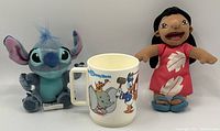 Front view of Stitch plush, vintage Disney coffee mug, and Lilo plush toy in a row.
