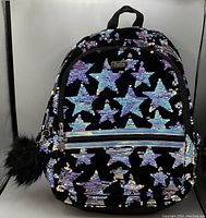 Photo of a black backpack with sparkly sequined stars in purple and blue with a black fuzzy pom-pom keychain on the zipper.