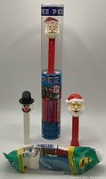 Photo showing five vintage PEZ dispensers: two Santa dispensers (one in original tube packaging with candies, one loose), one Snowman dispenser with black hat, one Bear dispenser in original plastic wrapper, and one extra Santa dispenser.