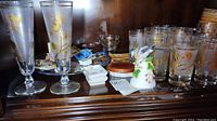 Top shelf display with two tall frosted beer glasses featuring gold pineapple and floral decorations, assorted small plates, pins and Wedgwood pin dish visible behind glasses.