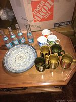 Overall view showing five vintage mugs in colorful floral and striped patterns, four Canada Dry sparkling water bottles, and a round ceramic serving tray on a wooden table