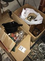 Two cardboard boxes of assorted kitchenware including plastic pots with handles, glass lids, metal utensils, and various other items.