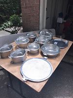 Full overview of 10-piece Guardian Service cookware set on a wooden table outdoors showing pots, pans, and lids with textured metal surfaces and glass lids.
