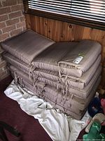 Stack of six beige outdoor cushions with tie straps, shown in an indoor setting against wood paneling and brick wall.