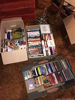 Three boxes of DVDs, CDs, and VHS tapes. VHS tape 'Field of Dreams' visible in one box. Multiple DVDs and CDs arranged in electronic media cases.