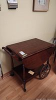Angles of a dark wooden tea cart showing drop leaf sides, two large wooden wheels, turned legs, and a lower shelf. Cart is on hardwood floor near wall and chair.