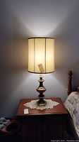 Single brass table lamp lit on wooden side table with cream colored fabric shade showing vertical texture and black trim edge. Lamp base is polished brass with turned design