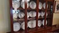 Plates and serving dishes arranged in a glass-front wooden cabinet, showing the floral porcelain pattern and gold trim.