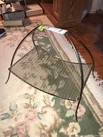 Side angled view of the metal magazine rack placed on a floral area rug near wooden furniture showing the curved legs and mesh metal tray.