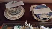 Two stacks of collector plates on a wooden surface, one with Norman Rockwell designs and the other with Royal Agricultural Society plates, some winter scenery shown on top plate.