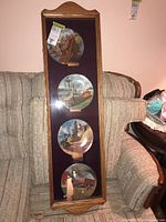 Four decorative plates displayed vertically in a wooden framed casing against a pink wall, on a beige upholstered couch, showing clear painted scenes on plates.