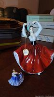 Two Royal Doulton figurines on a wooden surface, one larger in red dress and another smaller in blue.