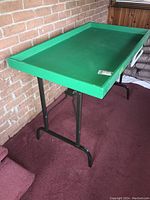 Side angle view of green folding table with black metal legs and wood top, placed near brick wall.