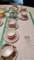 Full view of all nine teacups and saucers arranged in a line on a table with quilted white cloth.