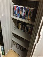 Five shelves inside a white cabinet filled with assorted VHS movies, showing multiple titles and some unopened packages.