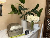 Wide view of large artificial plant, white vase with hydrangeas, and decorative metal chest with measuring tape for scale.