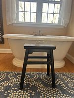 Black gloss Asian-inspired wooden stool placed in front of a white freestanding tub on a patterned rug. Visible light wear on stool corners.