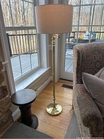 Full lamp showing brass finish, round base, tall slender pole, tan linen shade, and location near window and chair.