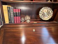 Full view of shelf with books, glass bubble weights, decorative plate with stand all on one wooden shelf inside secretary furniture