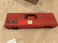 Closed view of red Sears Craftsman Large Truck Box on carpeted floor showing top side with stickers and handle.