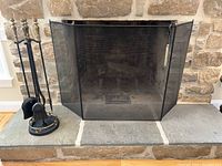 Photo of black mesh three-panel fireplace screen with brass corners and edge accents, placed in front of stone fireplace.