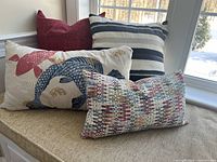 Four throw pillows placed on a window bench showing red lumbar, blue/cream striped, koi fish pattern, and multi-color patterned pillows