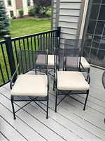Four matte black wrought iron outdoor chairs on a gray wood deck, each with beige cushions tied to the seats.