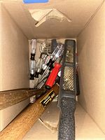 Top view into cardboard box showing two wooden-handled hammers, metal file with black handle, and several screwdrivers with clear and red handles.