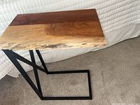 Full view of the C-shaped table showing the live-edge wood top and matte black metal frame alongside a bed.