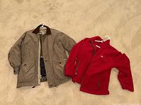 Two jackets laid on carpet floor; beige heavy duty Cabela jacket and red Gill polar fleece jacket side by side.
