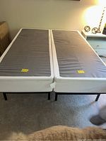 Photo showing the two metal box spring foundations placed side by side, covered in gray and white fabric with yellow tags, resting on carpeted floor.