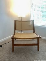 Front view of rattan chair showing woven rattan seat and backrest framed in solid wood with warm brown finish, situated in carpeted room next to lamp.