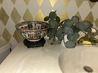 Wide shot of Rose Medallion bowl on carved wooden stand next to artificial plant on bathroom counter.